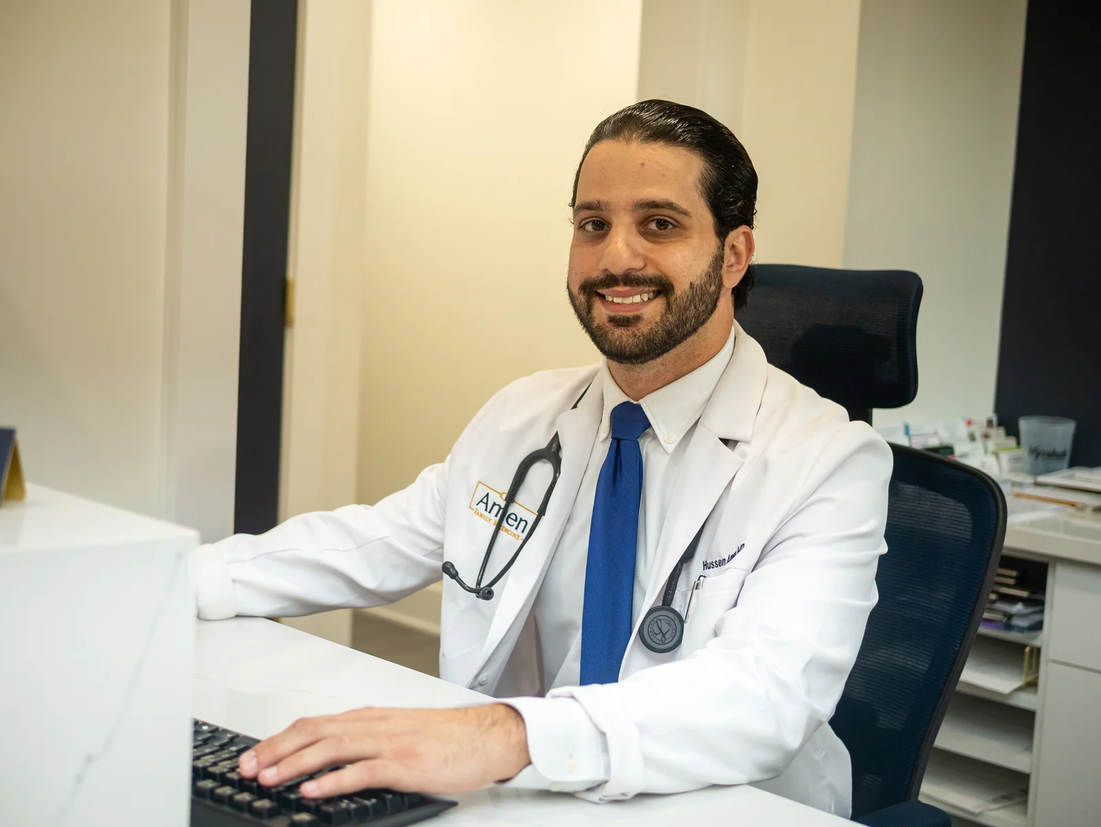 Dr. Hussen Amen smiling at his desk in a white lab coat with a stethoscope at Amen Family Medicine in Livonia.