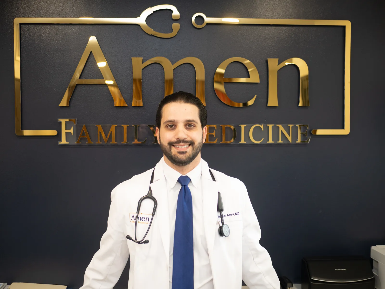 Dr. Hussen Amen, MD, board-certified family medicine physician standing in front of the Amen Family Medicine logo wall.